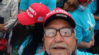 A man in a MAGA hat looks at the camera while taking a selfie in a crowd