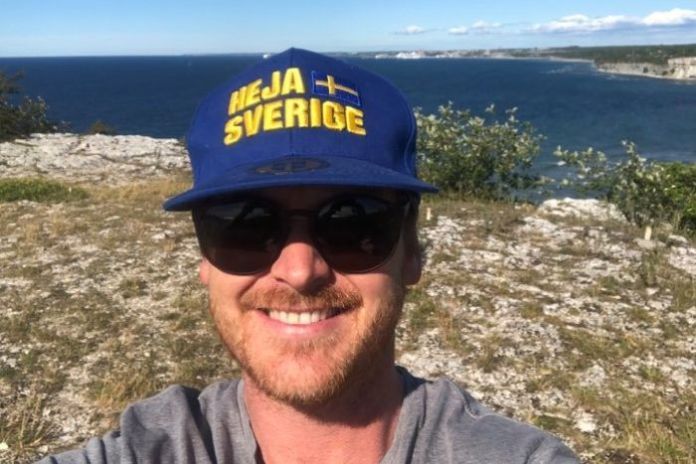 A man taking a selfie on the edge of a cliff wearing a blue and yellow hat with the Swedish flag on it.