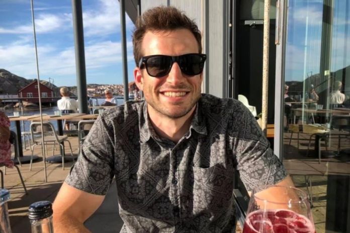Simon Keane, who wears sunglasses, is sitting at an outside table in a restaurant on a sunny day, having a drink in front of him.