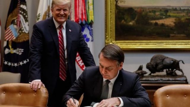 Trump smiling, standing behind the chair where Bolsonaro is sitting signing a document