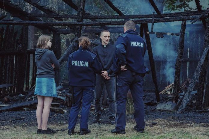 A scene from the film Corpus Christi with a young priest speaking to the police in front of a burned building