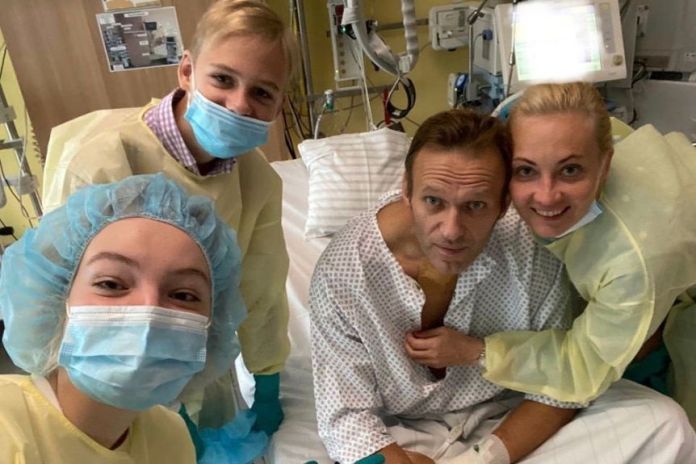 Alexei Navalny sits in a hospital bed in a medical gown, surrounded by three people in protective gowns, masks and hair nets