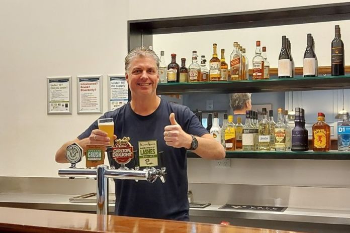 A man stands behind a bar smiling and holds a beer in his hand.
