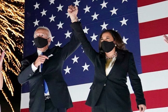 Joe Biden and Kamala Harris raise their hands together in front of a US flag as fireworks explode behind them