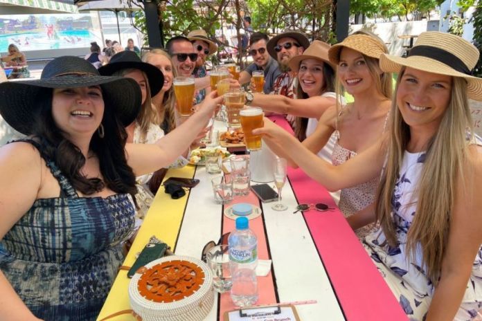 A group of 10 friends sit at a long table and lift their glasses.