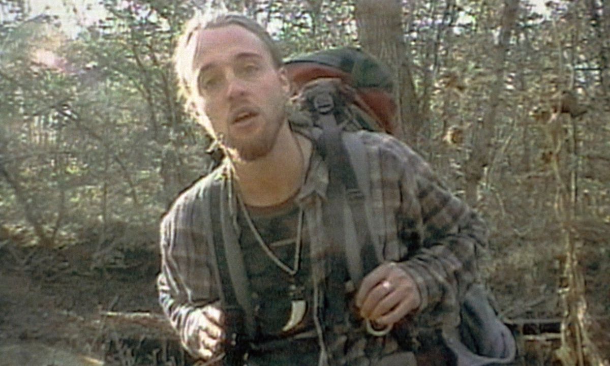Joshua Leonard stands in a forest in a dingy flannel and heavy backpack, staring at the camera in The Blair Witch Project.