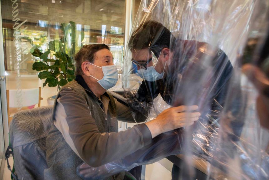 Relatives hug each other through a plastic wrap and glass while wearing masks.