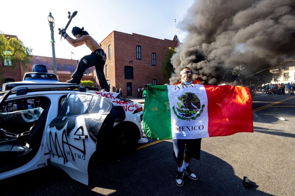 A demonstrator strikes a Waymo self-driving car with a skateboard as another burns in the background during a protest against federal immigration sweeps in downtown Los Angeles June 8, 2025. — Reuters pic